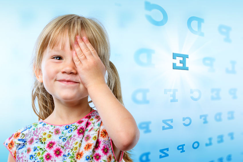 An image of a young girl smiling at the camera whilst holding one of her hands over her left eye. In the background there is a combination of letters, similar to those you would see during an eye test.