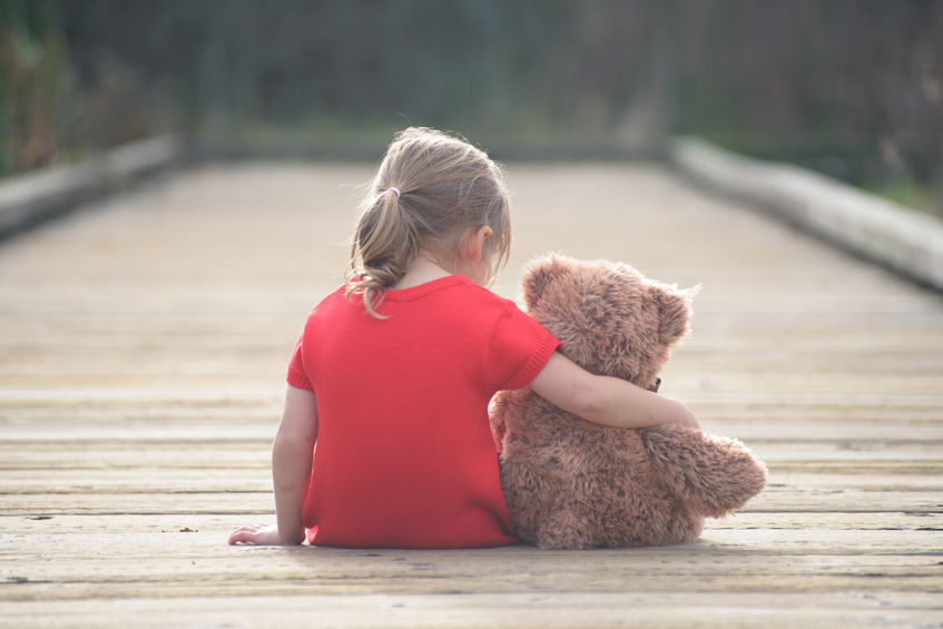 A child sat down outside on some decking with their back to the camera. They have their arm wrapped around a teddy bear.