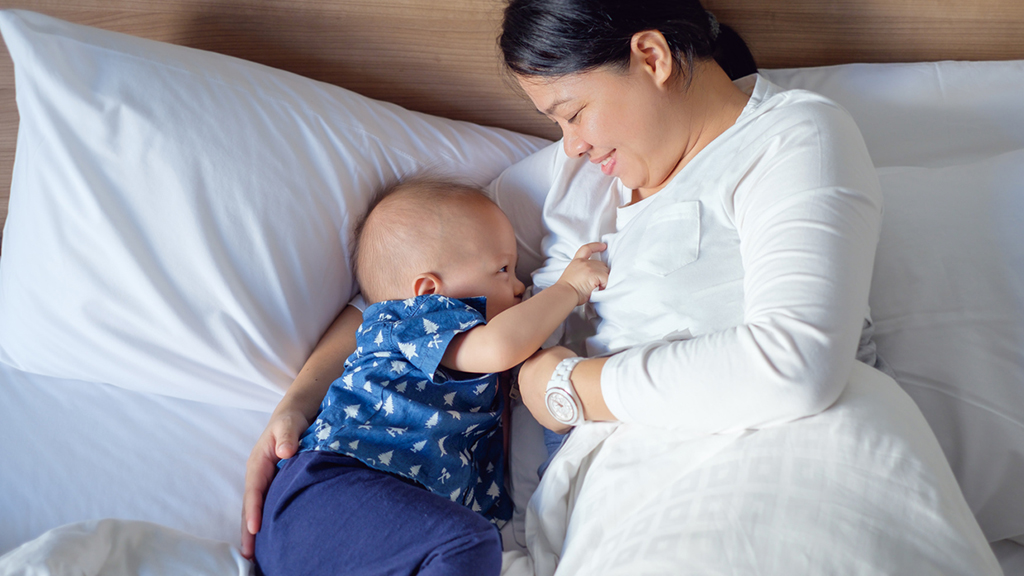 Mother and baby re both lying down in bed together. They are lying down on their sides and facing each other - the mother has her arm positioned behind the baby to support them.