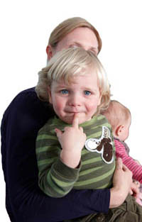 Blonde, male toddler looking at the camera and pointing at his mouth. There is a woman positioned behind him holding a baby - presumably his mother and sibling.