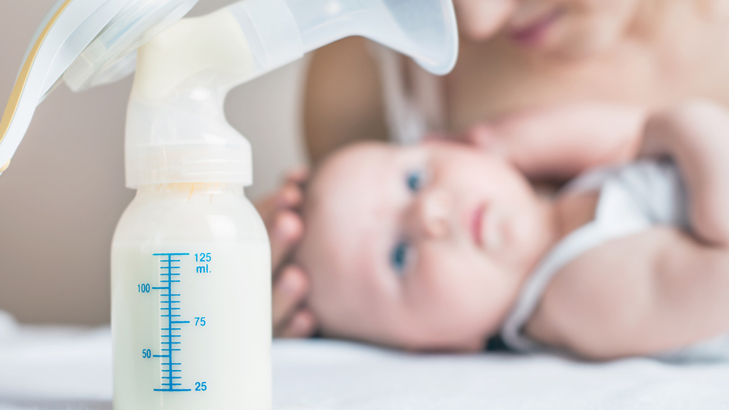 An image of a baby bottle that contains colostrum (first milk). In the background of the image there is a baby lying down out of focus.
