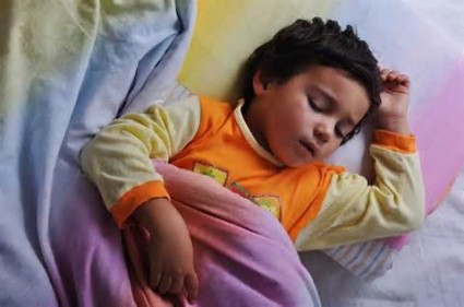 A young child asleep in their bed - they are lying down on their back with their arms above their head.