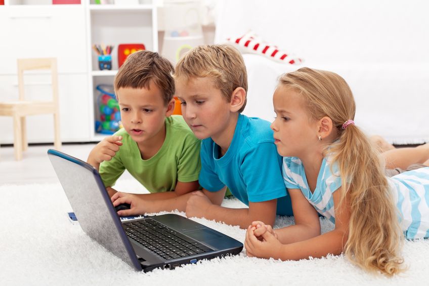 Three young children - two boys and one girl - lay down on the floor in a bedroom, looking at a laptop screen.