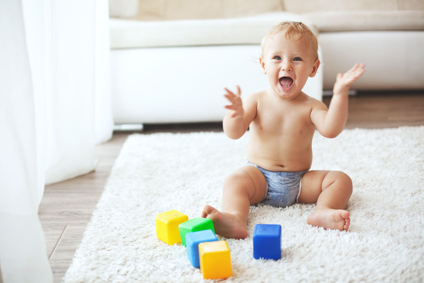 Male toddler sat on a rug, waving his arms in the air excitedly whilst looking at the camera. There is a selection of toy building blocks by his feet.