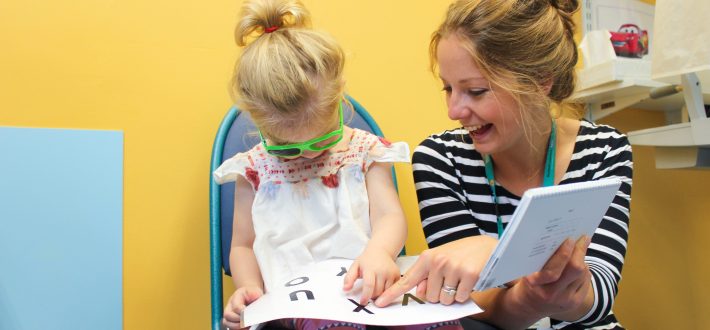 Female optician doing an eye test with a female toddler. The woman is pointing at a sheet of letters whilst the toddler attempts to read them whilst wearing a pair of glasses.