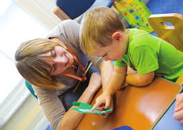 A young child and a teacher sat at a table together. The child is pointing at some words on a piece of paper whilst the teacher looks on encouragingly 