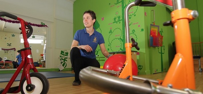 A white, female, presumably nursery nurse, knelt down in the middle of the nursey floor whilst surrounded by different pieces of apparatus and equipment e.g. a giant bouncy ball and a scooter.