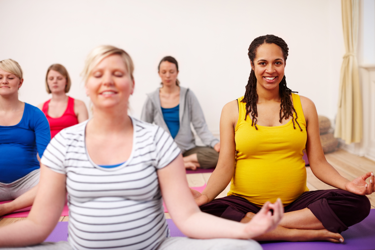 An image of 5 expectant, pregnant females sat down on matts doing yoga. They are sat in a cross legged position - one woman is looking at the camera and smiling whilst the other 4 women have their eyes closed. cross legged